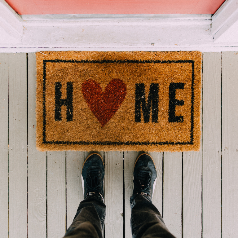 door matt with word home, red love heart insead of the o in home. person stood in front of matt with black trousers and shoes. you can only see legs and feet