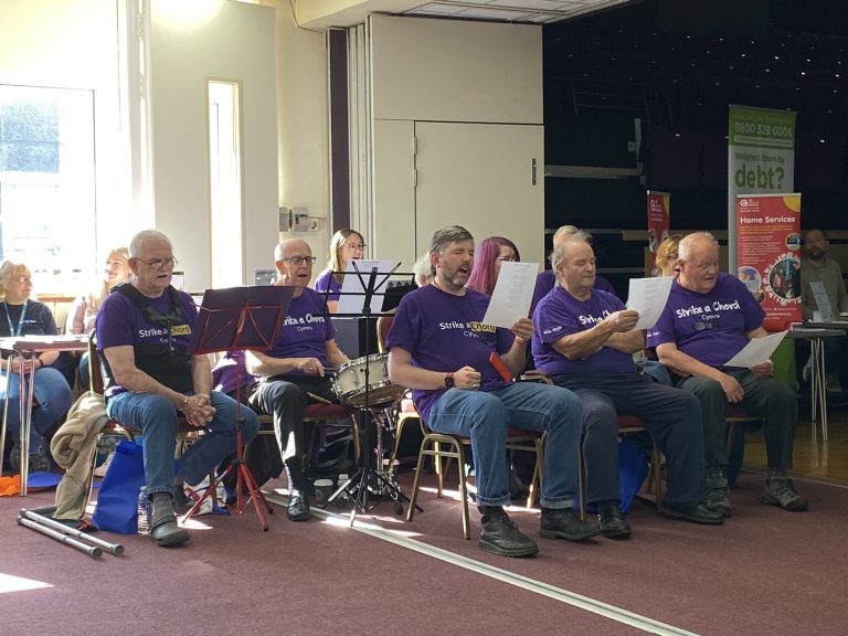 men sat down, some with instruments ready to sing, wearing purple Stroke Association tshirts with Strike a Chord written in white